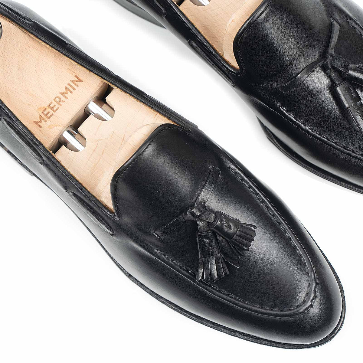 Close-up top view of Meermin black boxcalf tassel loafers on wooden shoe trees, showing handsewn apron and hand-closed tassel detail