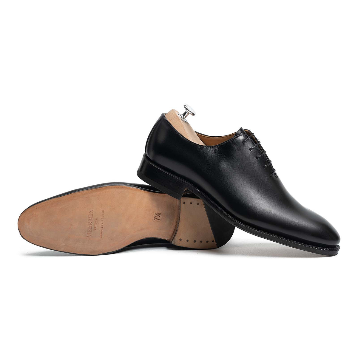 Meermin black boxcalf wholecut Oxford pair, one shoe on shoe tree showing side profile, the other resting sole-up revealing leather sole