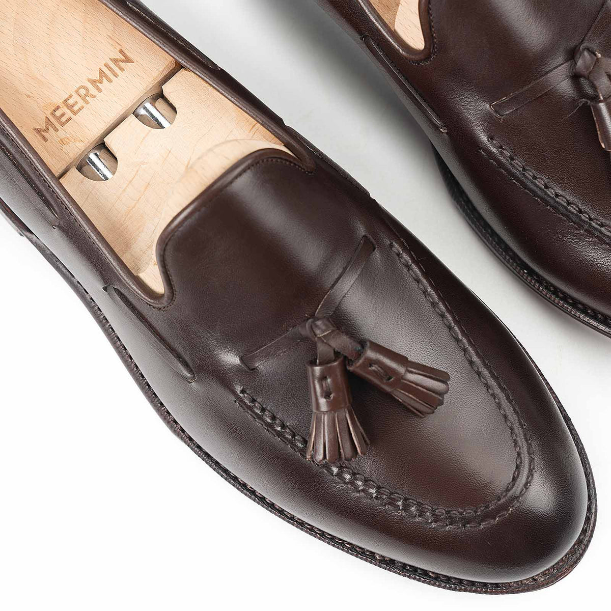 Close-up of Meermin dark brown boxcalf tassel loafers on wooden shoe trees, showing hand-closed tassels and apron stitching detail