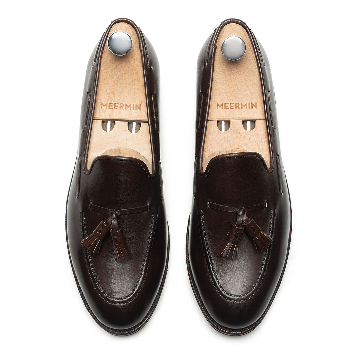 Meermin dark brown boxcalf tassel loafers, top-down view on shoe trees, showing hand-closed tassels and apron stitching