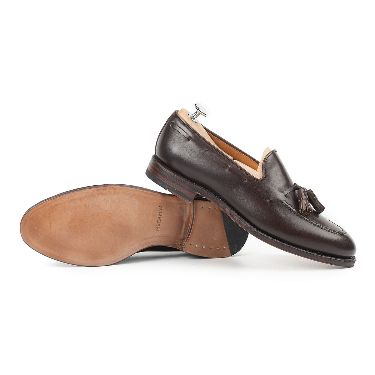 Meermin dark brown boxcalf tassel loafers, one shoe on a wooden shoe tree, the other resting sole-up to show the leather sole and heel stack