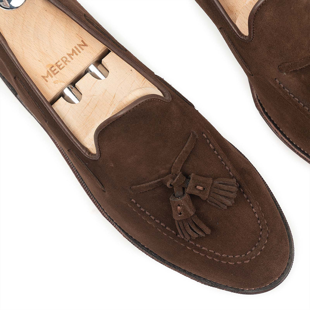 Close-up of Meermin brown suede tassel loafers on shoe trees, showing hand-sewn apron stitching and tasseled vamp detail