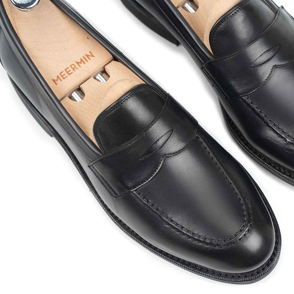 Close-up of Meermin black silkcalf penny loafers on wooden shoe trees, showing the apron stitching and rounded toe detail