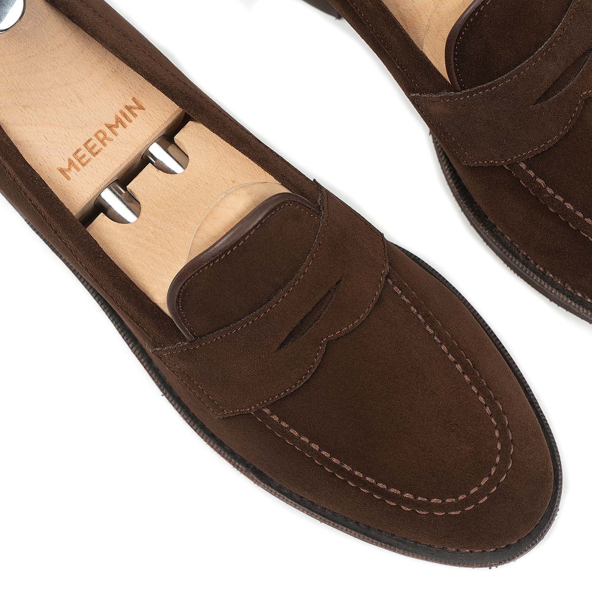 Close-up of Meermin brown suede penny loafers on wooden shoe trees, showing saddle stitching and diamond latex rubber sole detail