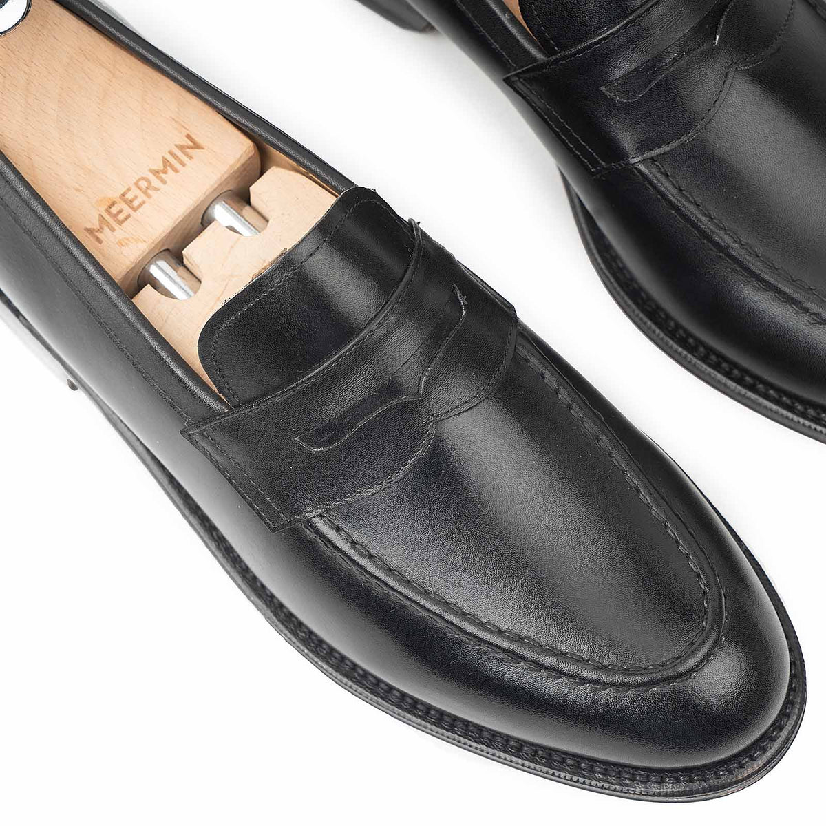 Close-up top view of Meermin black boxcalf penny loafers, one fitted with a wooden shoe tree, showing saddle strap detail