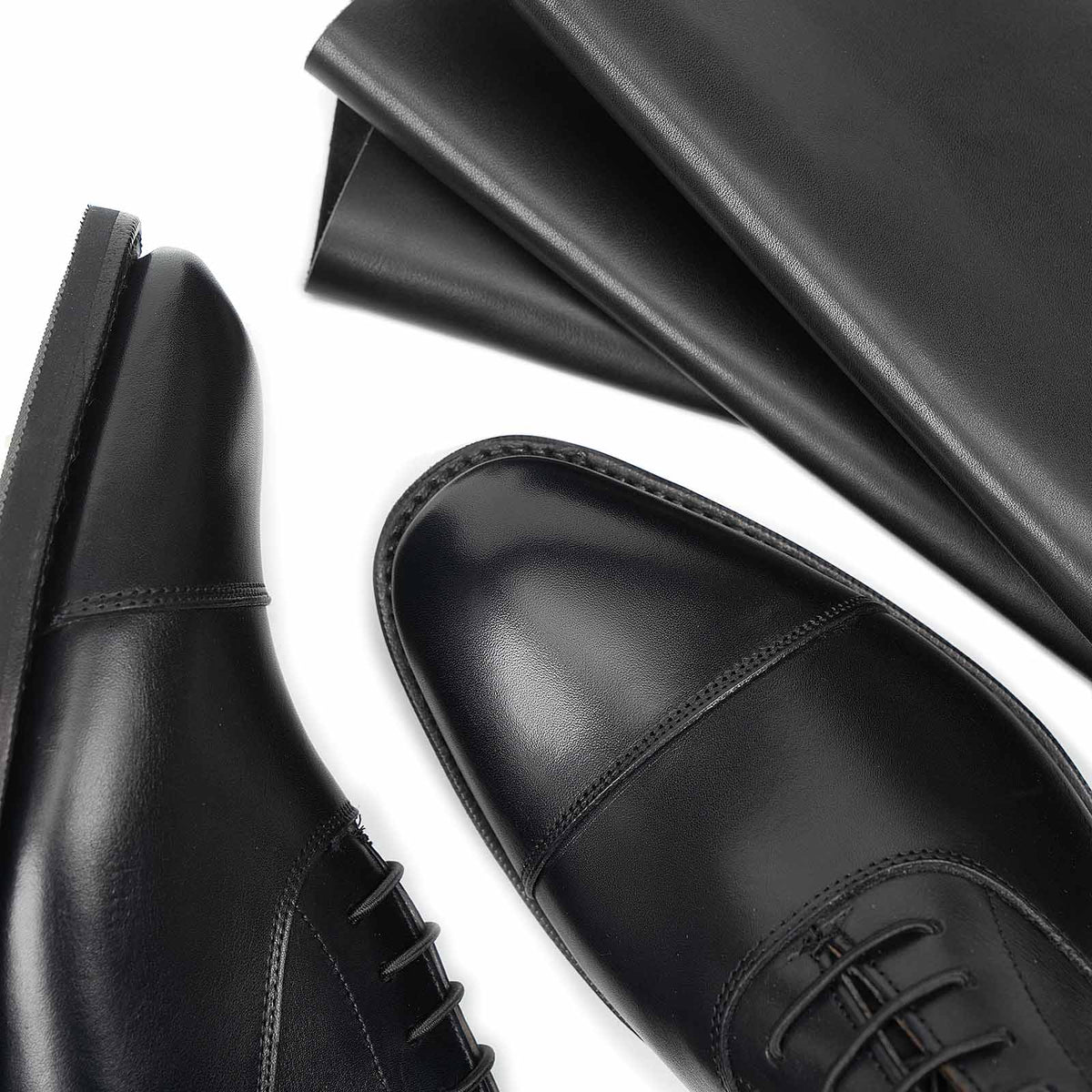 Meermin black silkcalf cap-toe Oxfords, close-up on toe caps and stitching beside a folded sheet of the raw leather