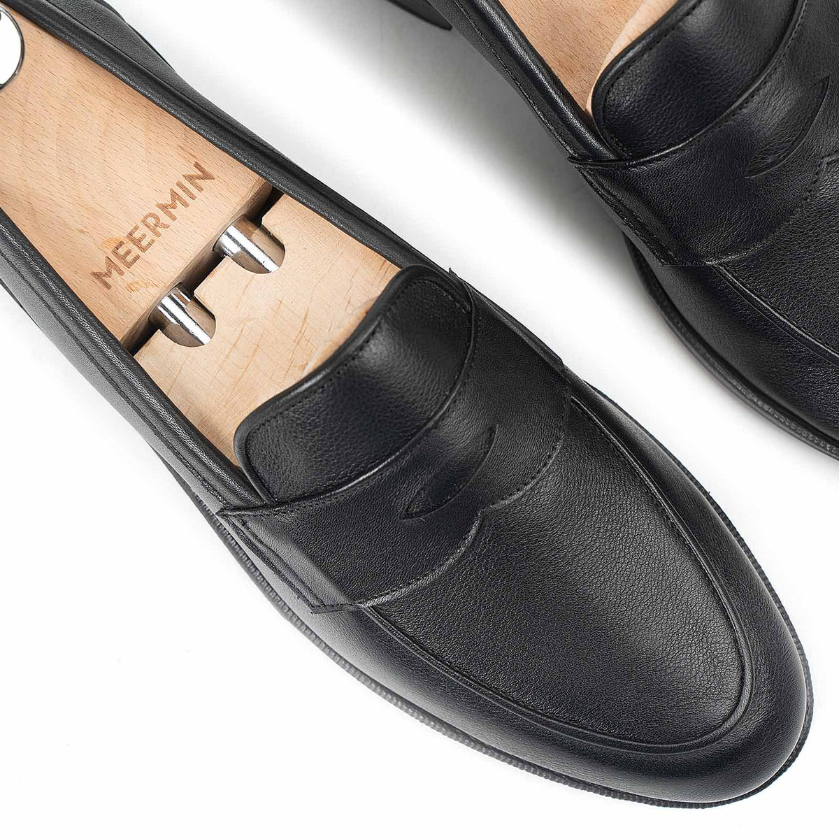 Close-up top view of Meermin black softcalf penny loafers on wooden shoe trees, showing the apron piping and moc-toe seam detail