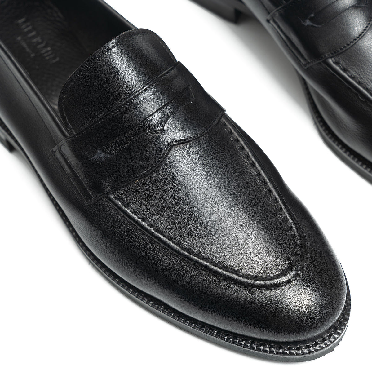 Close-up top view of Meermin black softcalf penny loafers, showing the saddle strap, apron stitching, and round toe detail