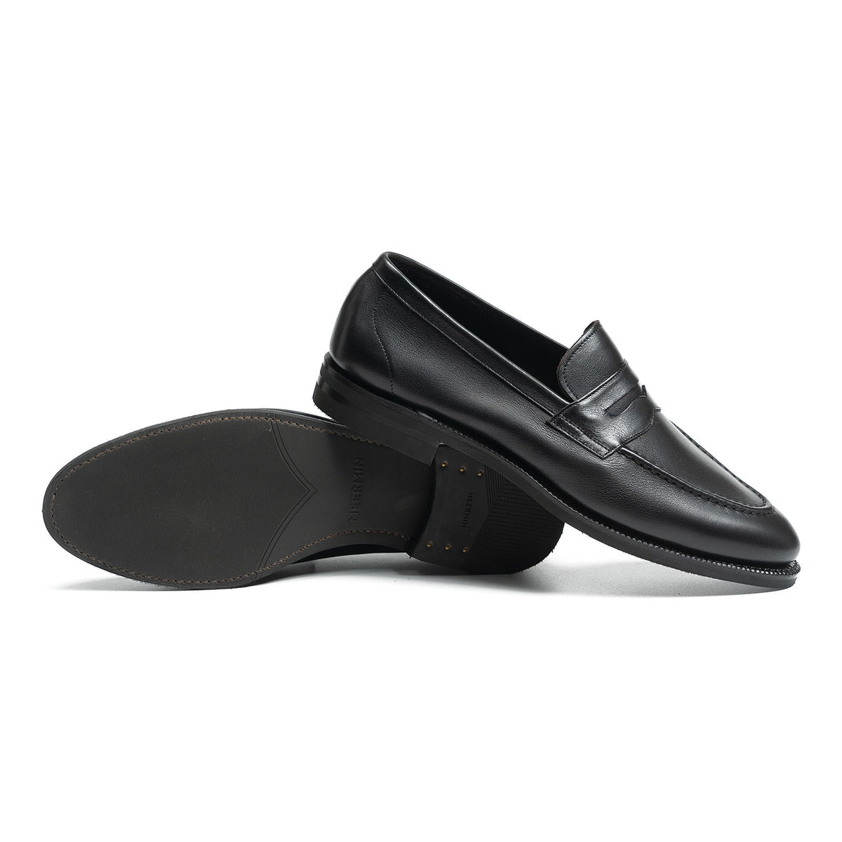 Meermin black softcalf penny loafers shown paired, one resting on its side atop the other to reveal city rubber sole detail