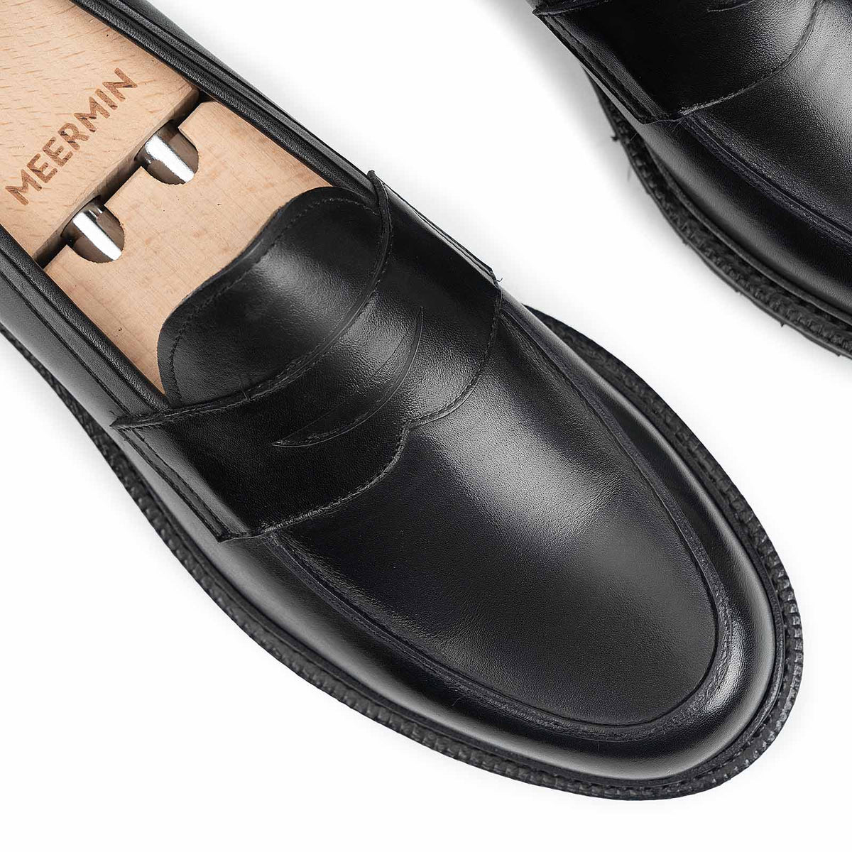 Close-up of Meermin black boxcalf penny loafers on a wooden shoe tree, highlighting the stitched apron and cleated rubber sole